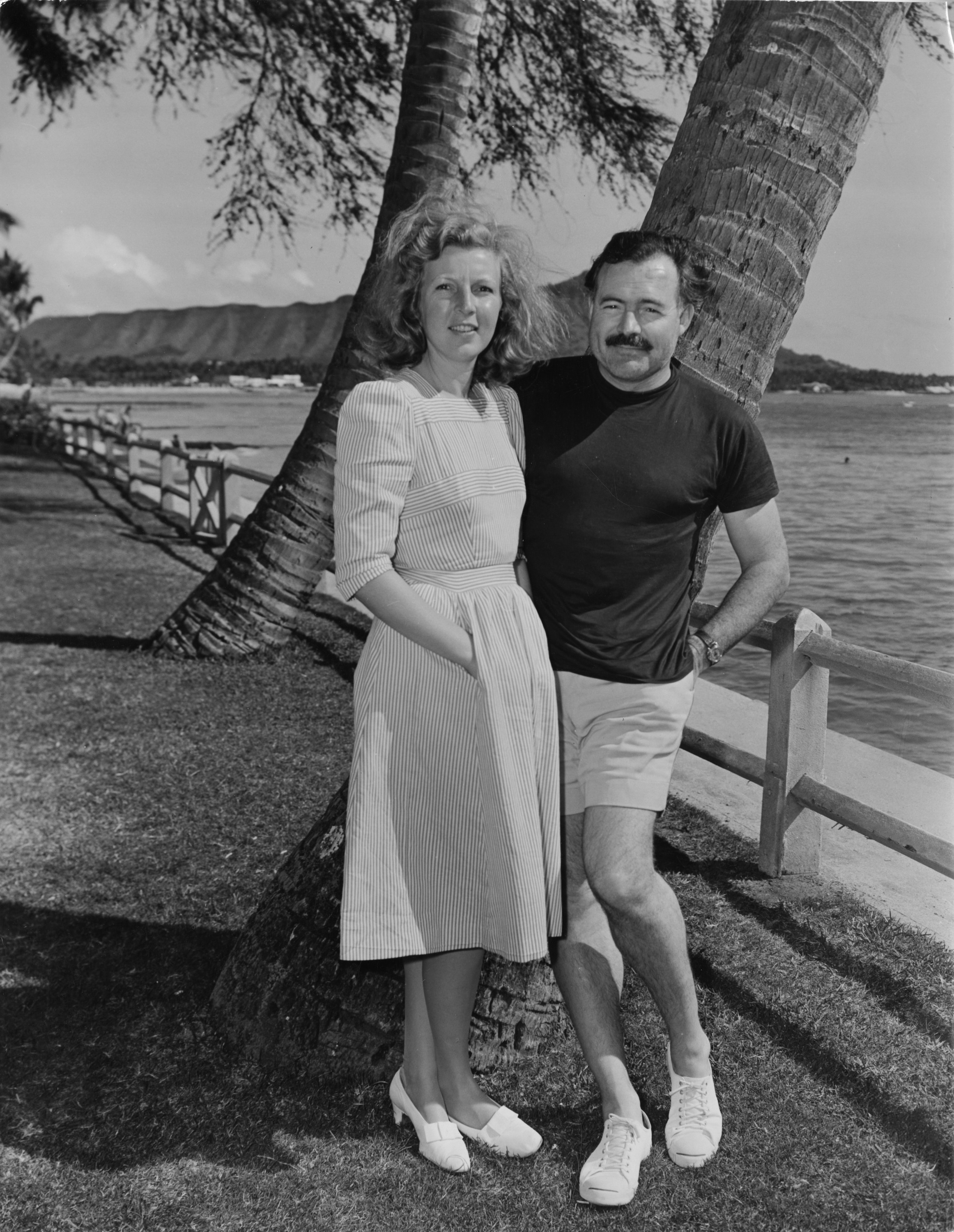 Black and white image of Ernest Hemingway, wearing a dark t-shirt, light shorts, and sneakers, and Martha Gellhorn, wearing a light striped dress and low-heeled light shoes, leaning against a palm trunk by the ocean.