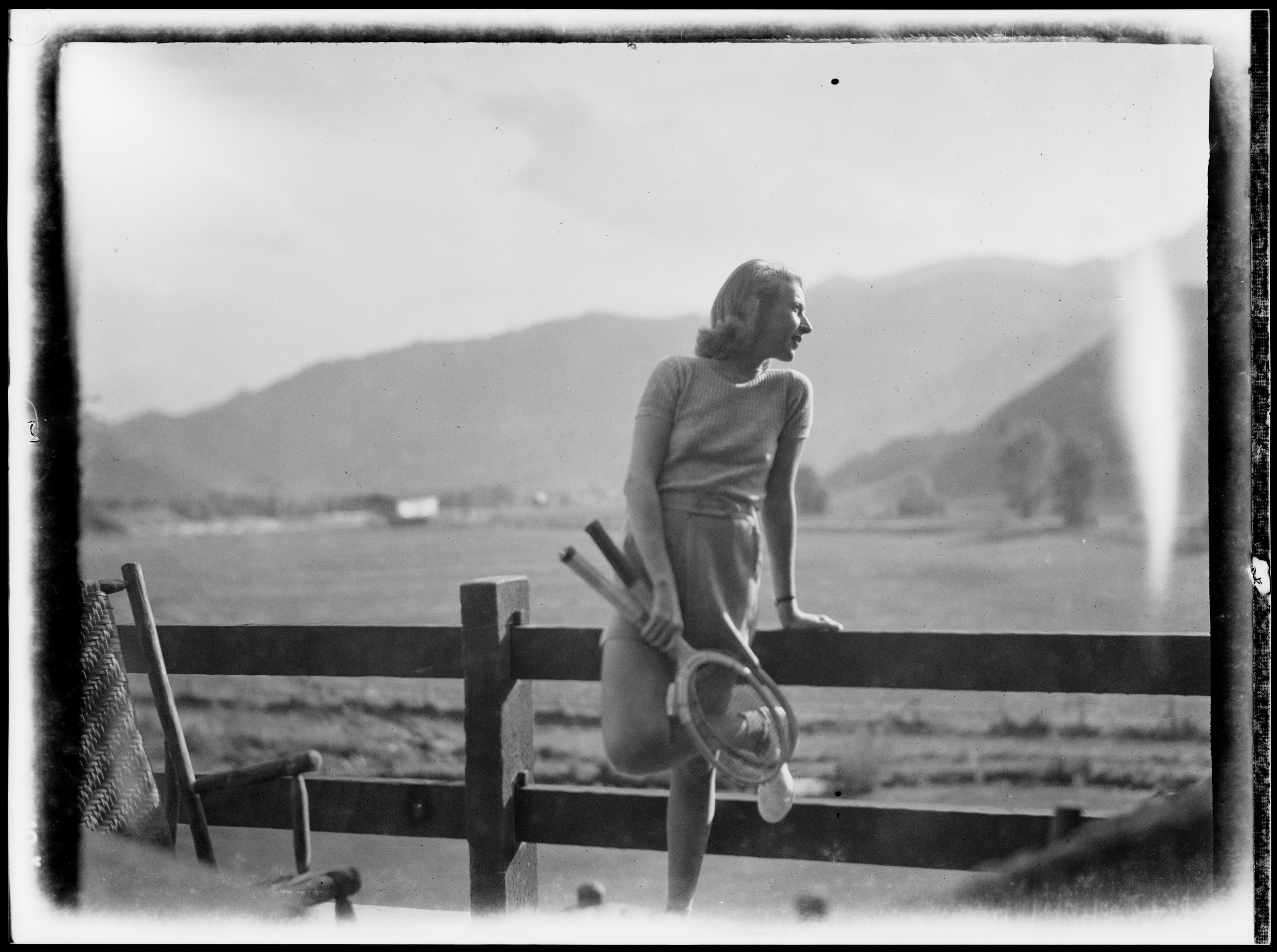 A black and white image of Martha Gellhorn leading against a fence in Sun Valley Idaho.  She holds two tennis rackets and looks away to the right, her face catching the sun.  Behind her is a flat field and, farther away, mountains.