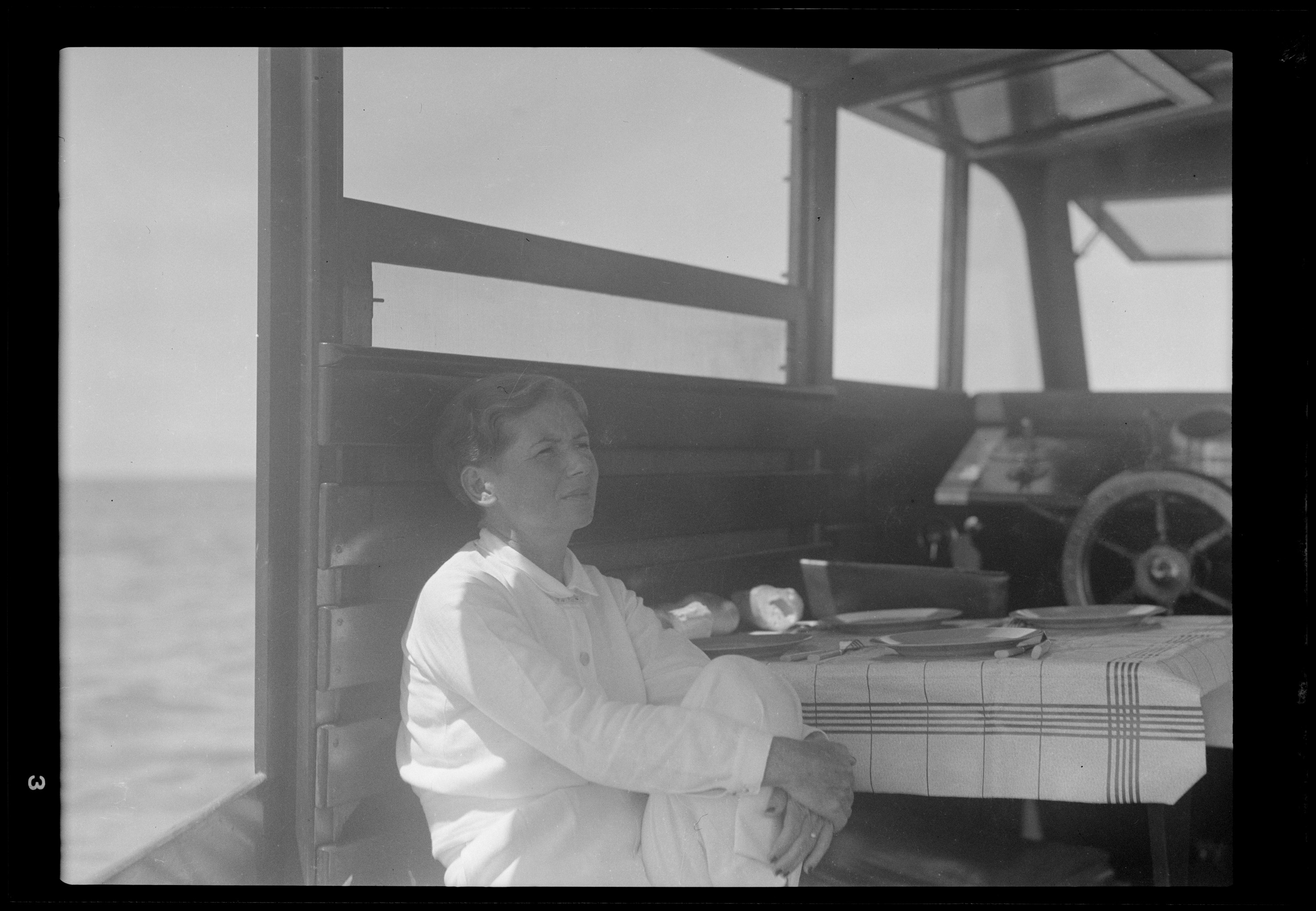 A black and white image.  Pauline Hemingway sits in the cabin of the Pilar, next to a table covered with a checked cloth.  She wears a white long sleeved shirt, white pants, and light-colored earrings.  Her knee is drawn up and clasped loosely in her arms.  She gazes off toward the right; the sea is visible behind her.
