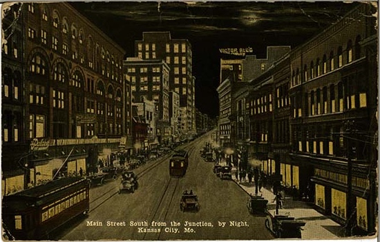 The front of a postcard showing "Main Street South from the Junction by Night / Kansas City, MO." A street scene showing multi-story buildings, mostly brick, from four to eight stories high.  Cars are parked along the sidewalk to the right; in the center and on the left side of the street, trolley cars and tracks.  The sky is dark, with a moon and wispy clouds.  The scene is lit by store-front windows, auto and trolly car headlights, and lights in the upper-story windows of the buildings.