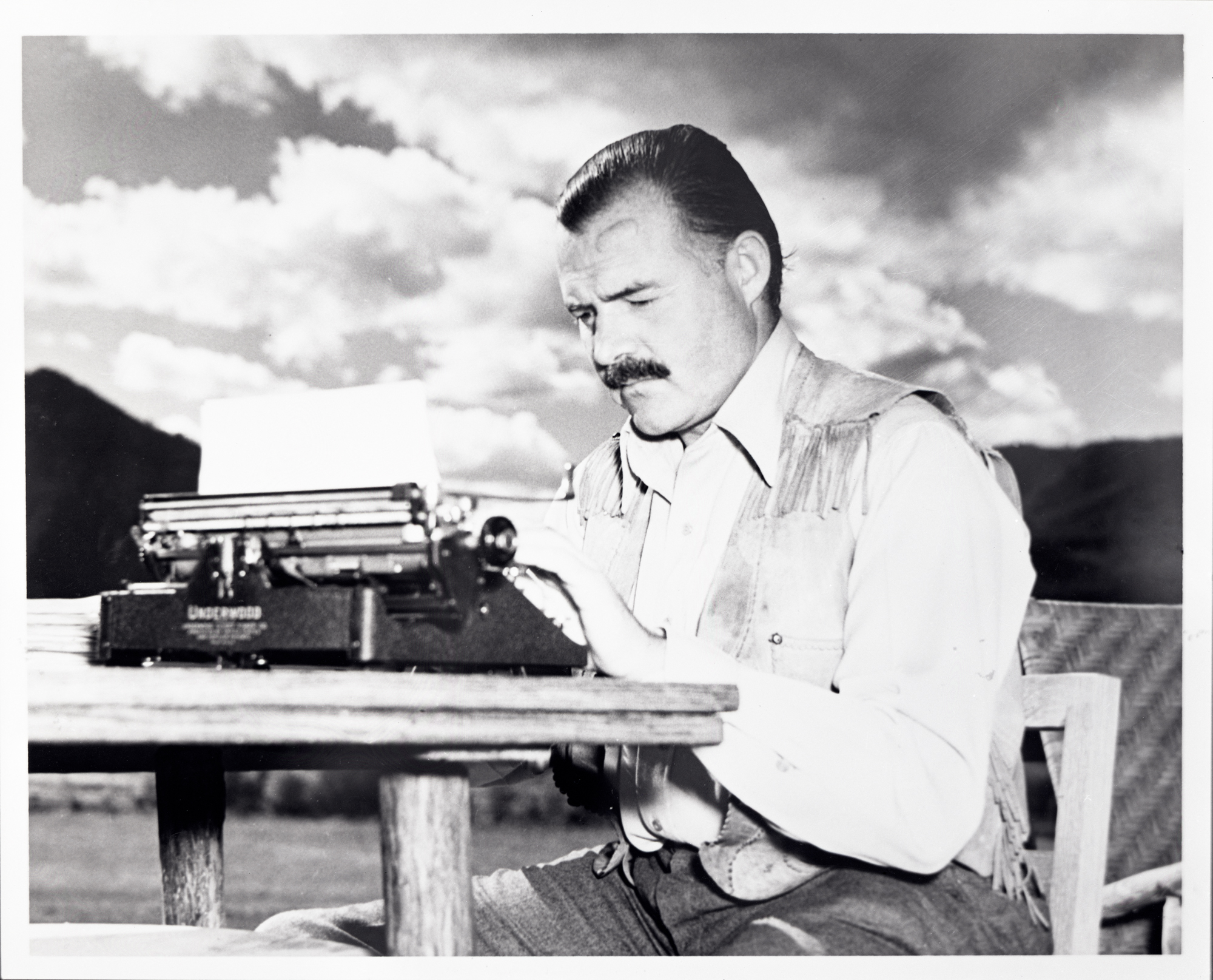 A posed black and white photograph of Ernest Hemingway typing outdoors. He wears jeans, a white shite, and a fringed vest. Behind him are mountains and a dramatically cloudy sky. He sits at a hewn wooden table.