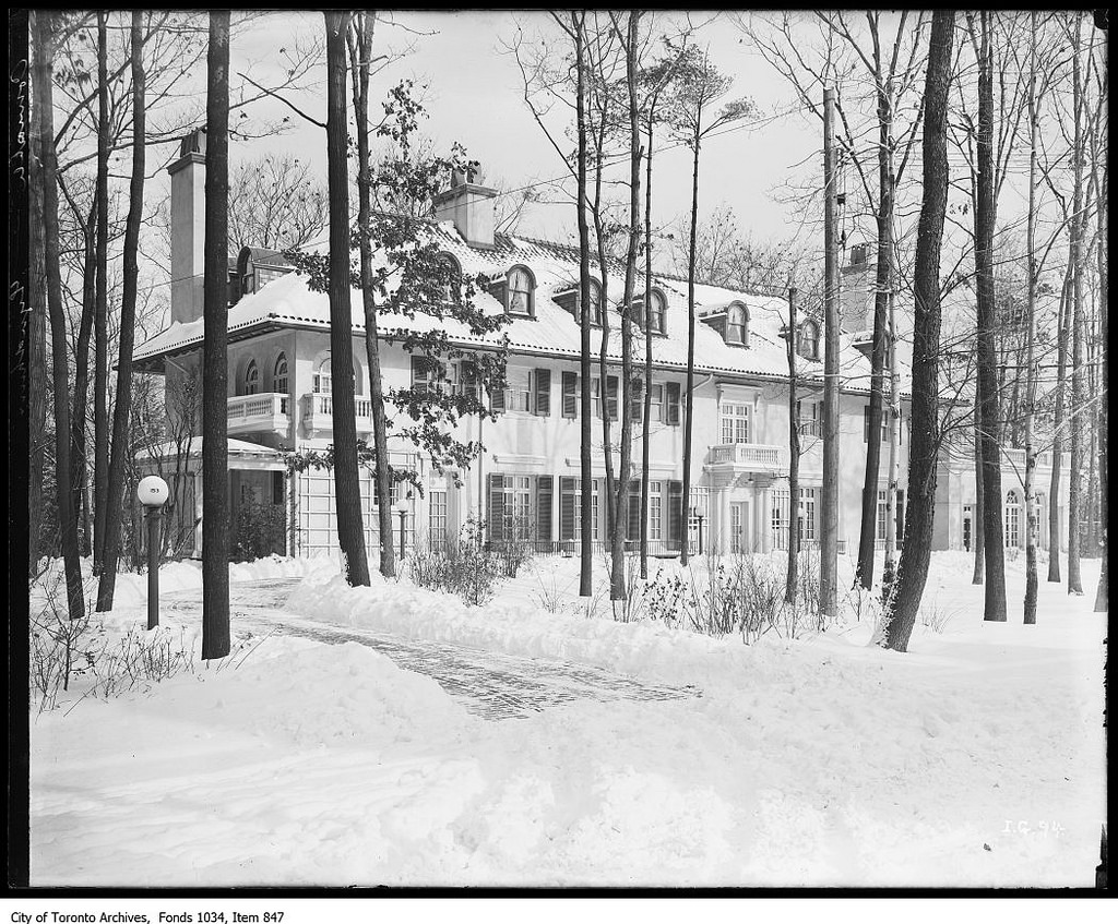 A black and white photograph of a large, stately home.  The home is long and white with a pillared central entrance. There is a covered carriage portico on one end.  The home is white, matching the deep snow in the wooded yard. The driveway has been plowed.