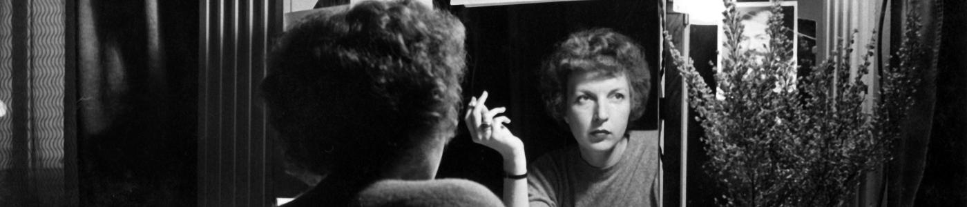 Martha Gellhorn reflected in a mirror, writing.  Photos of Ernest Hemingway hang on the mirror's frame.