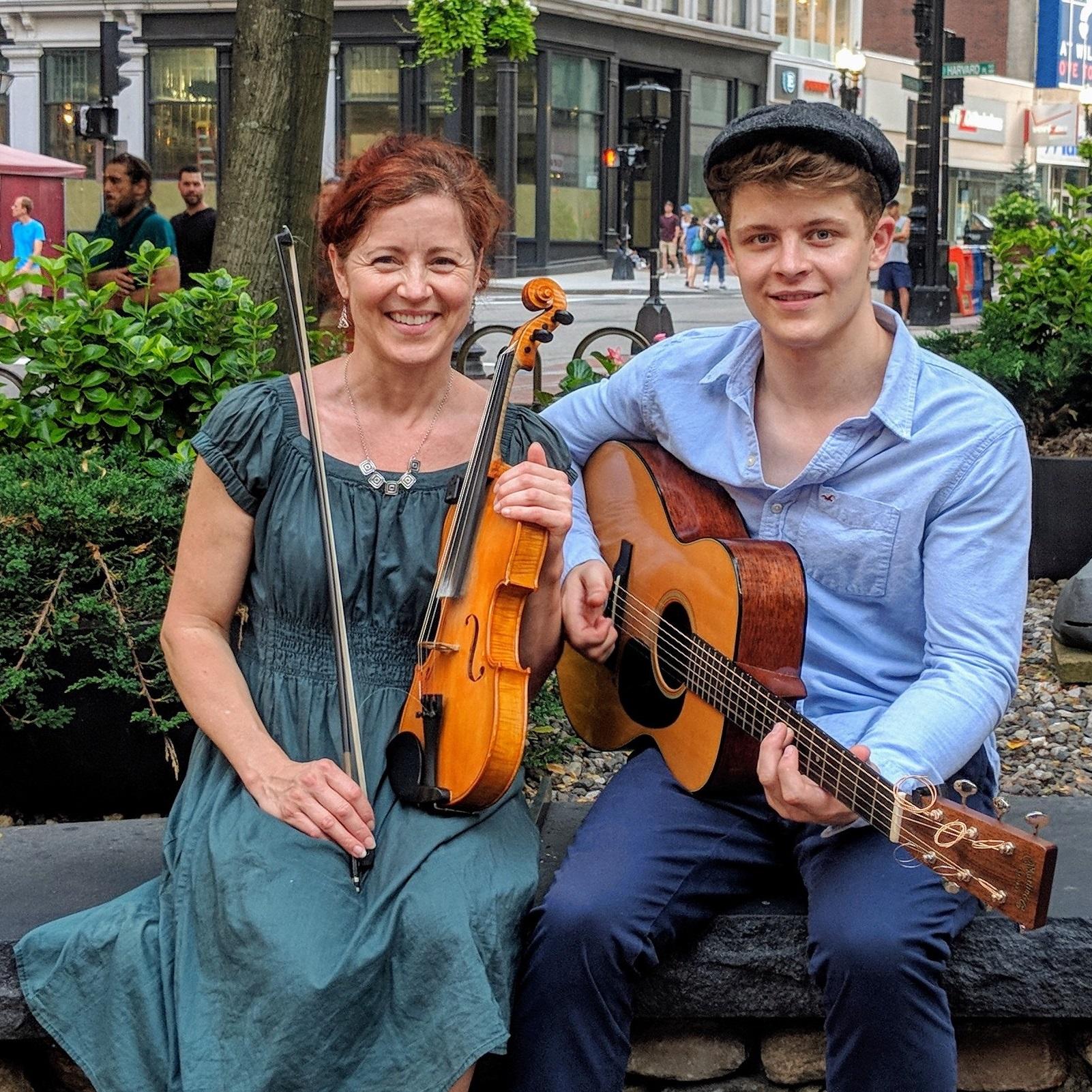 Image of Nancy Bell sitting next to Calum Bell, she is holding a violin and he is holding a guitar.