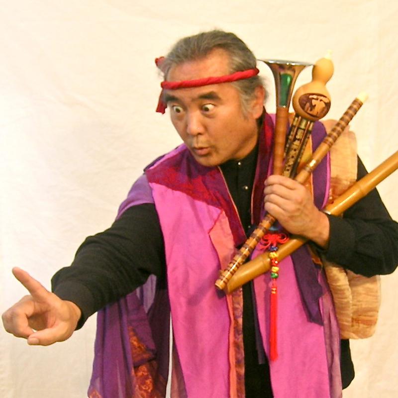 A man holding several wind instruments points amusingly. 