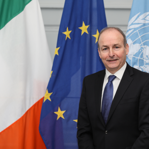 Micheál Martin stands facing the camera and smiling in front of three flags, including the flag of Ireland.