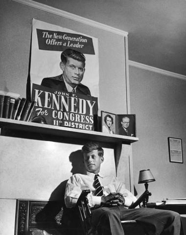 JFK seated under a Congressional campaign poster