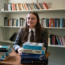 Ruby McIntee, an 18 year-old young woman with long brown hair parted down the middle is in a gray school uniform with a white shirt and striped tie. There are two stacks of books in front of her and shelves of books behind her.