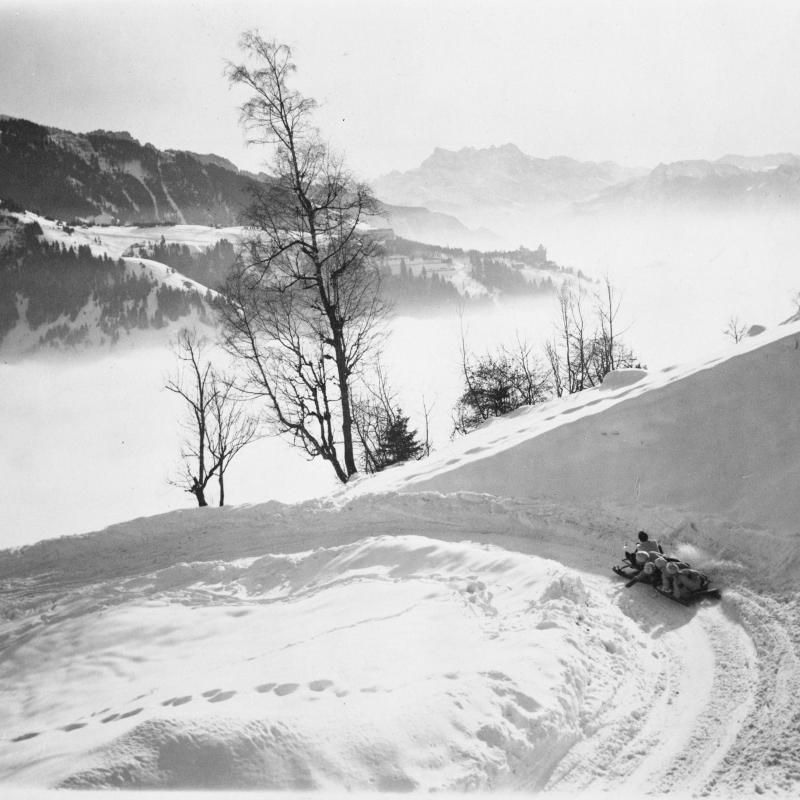 A black and white photograph of the Sonloup bobsled run in the Swiss Alps, near Montreux.  Hemingway with three others rides a bobsled into a curve on the run.  Mountains and mist in the background.