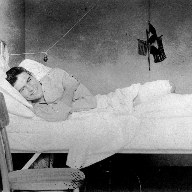 The 19-year-old Ernest Hemingway smiles from his hospital bed, Red Cross Hospital, Milan, 1918.  A small U.S. flag hangs on the wall behind him.