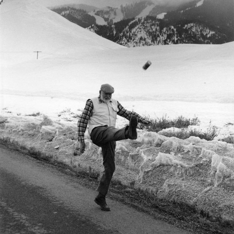 Ernest Hemingway kicks a can down the road in Ketchum, Idaho, c. 1960.  Snow-covered mountains with low clouds in the distance.