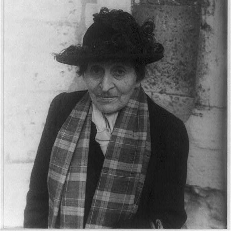Black and white portrait photograph of Alice B. Toklas, Chartes, 1949. Toklas wears a black hat decorated with feathers, a plaid scarf, a dark jacket, and a light-colored blouse. She is leaning against stonework and looks at the camera.