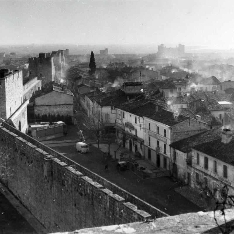 A black and white image taken from atop a rampart tower, thus a top-down view of a French walled town with the sea in the distance.  Stone ramparts punctuated by medieval defense towers wrap the town on the left side of the image.  Inside the walls are a street and a row of two or three story white buildings with dark roofs.  