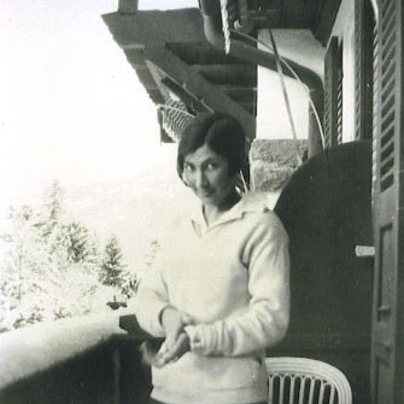 A black and white photographic image. A young woman dressed for skiing stands on a snowy balcony. She has angled cropped dark hair and, her head tilted down, she looks up at the camera.  A bentwood chair is behind her; over the balcony rail, snowy pine trees and mountains are just visible in the distance.