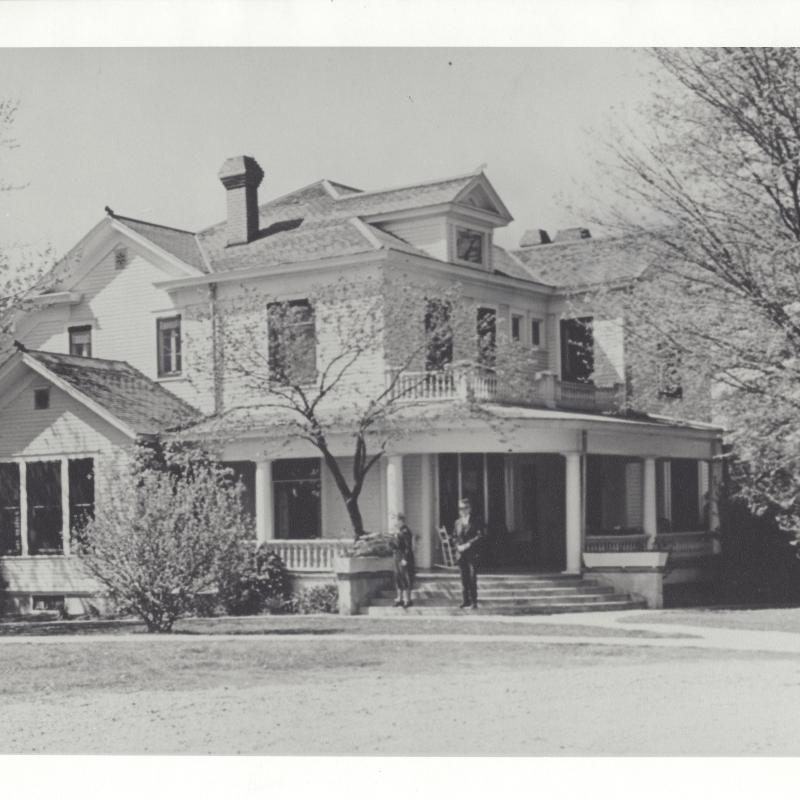 A c. 1930 black and white photograph of a white wooden frame house with a wraparound porch, the Pfeiffer family home in Piggott, Arkansas.  Paul and Mary Pfeiffer (Pauline Hemingway's parents) stand on the porch steps.