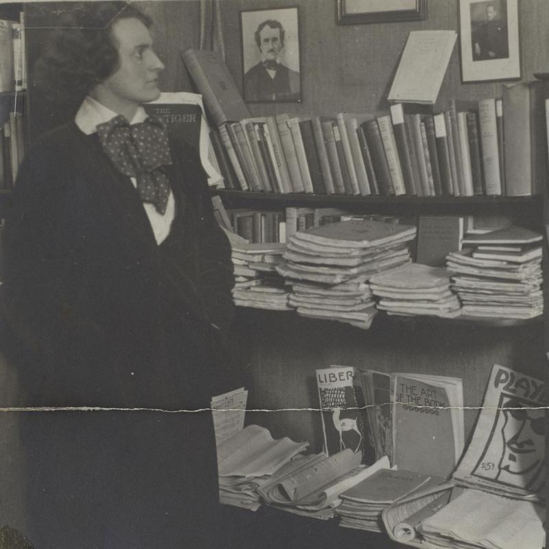Detail from a photographic black and white image affixed to an information card. The image shows Sylvia Beach, wearing a dark suit, white collared blouse, and a silk tie standing in front of the bookshelves in her bookshop.